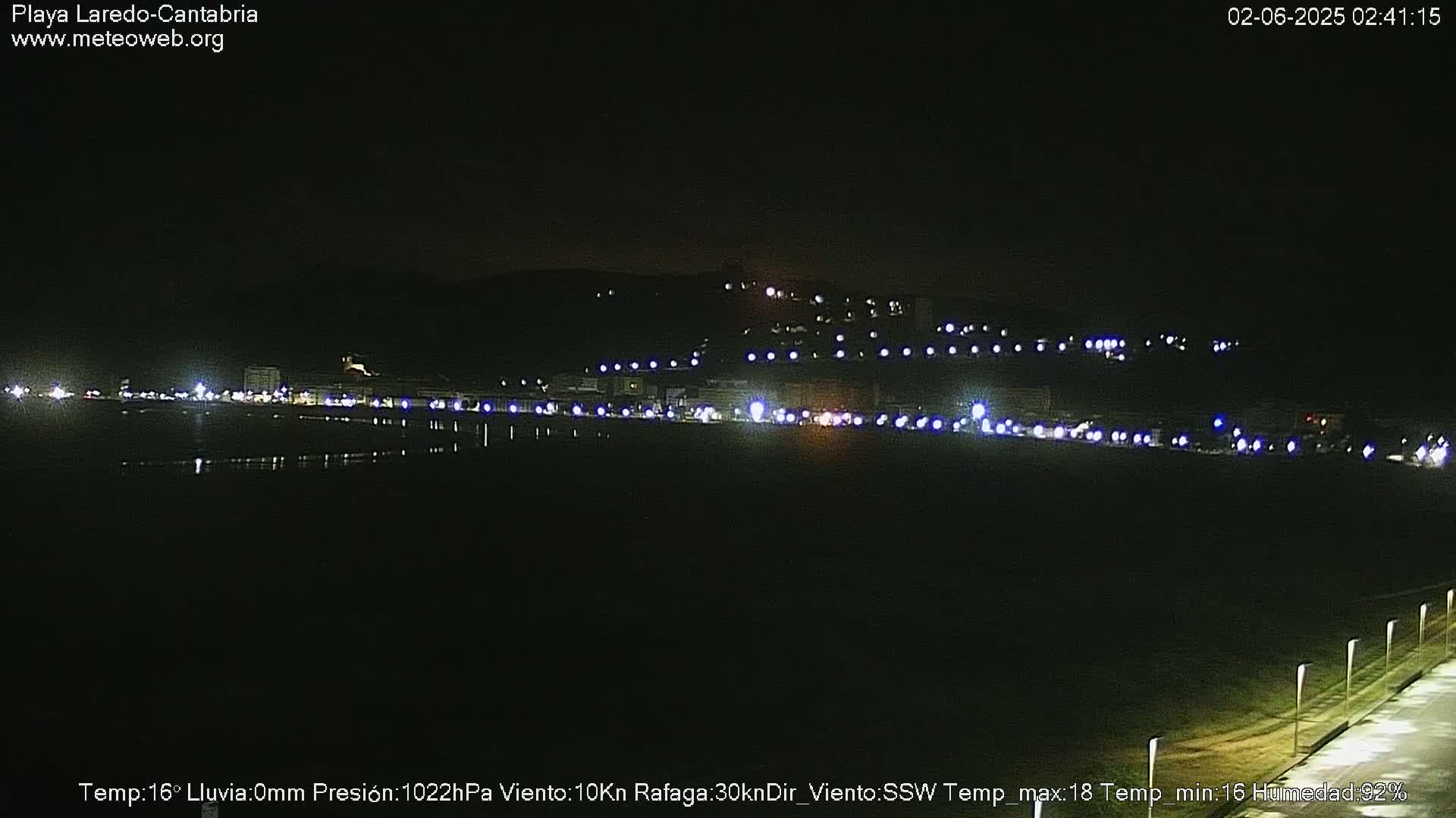 A nighttime view of a dark, flat beach with a line of lights from buildings and vehicles in the distance under a clear, dark sky.