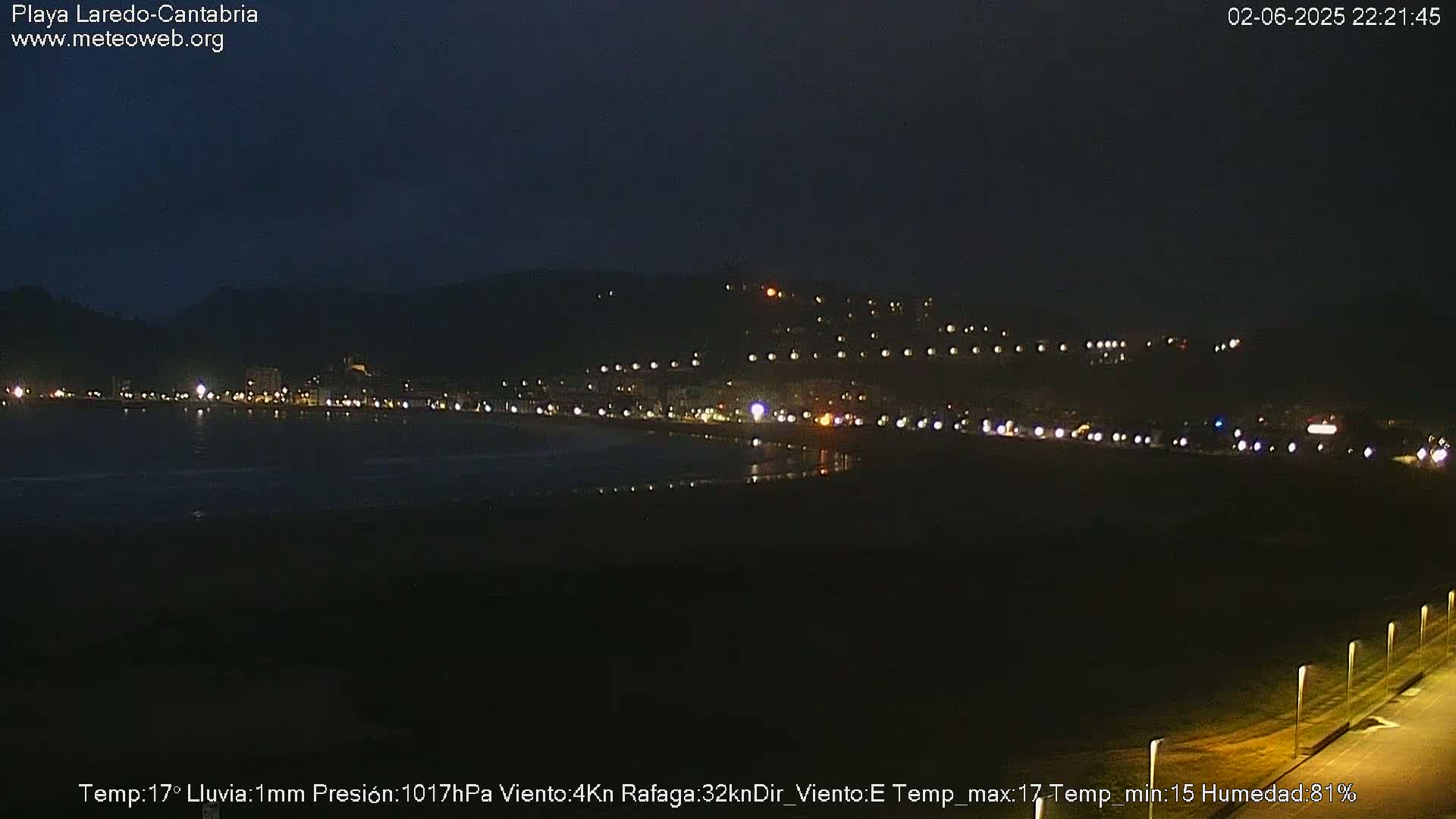 A nighttime view of a coastal town illuminated with lights, under a dark sky with light rain.