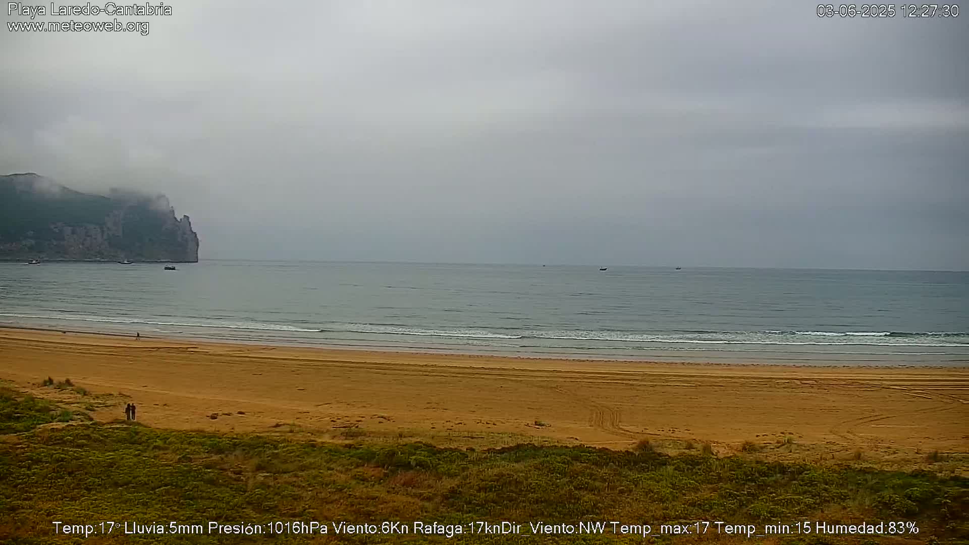 A wide sandy beach stretches to a calm, gray sea under an overcast sky, with a distant, fog-shrouded cliff visible in the background.