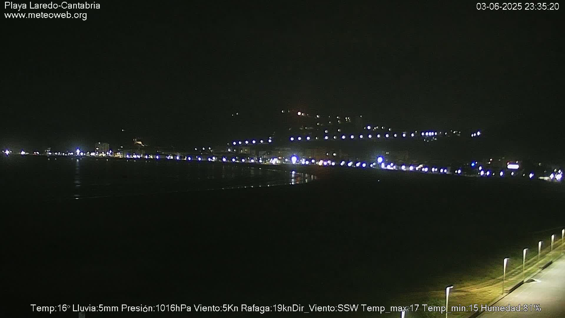 A nighttime view of a coastal town with numerous lights along a dark, wet shoreline.