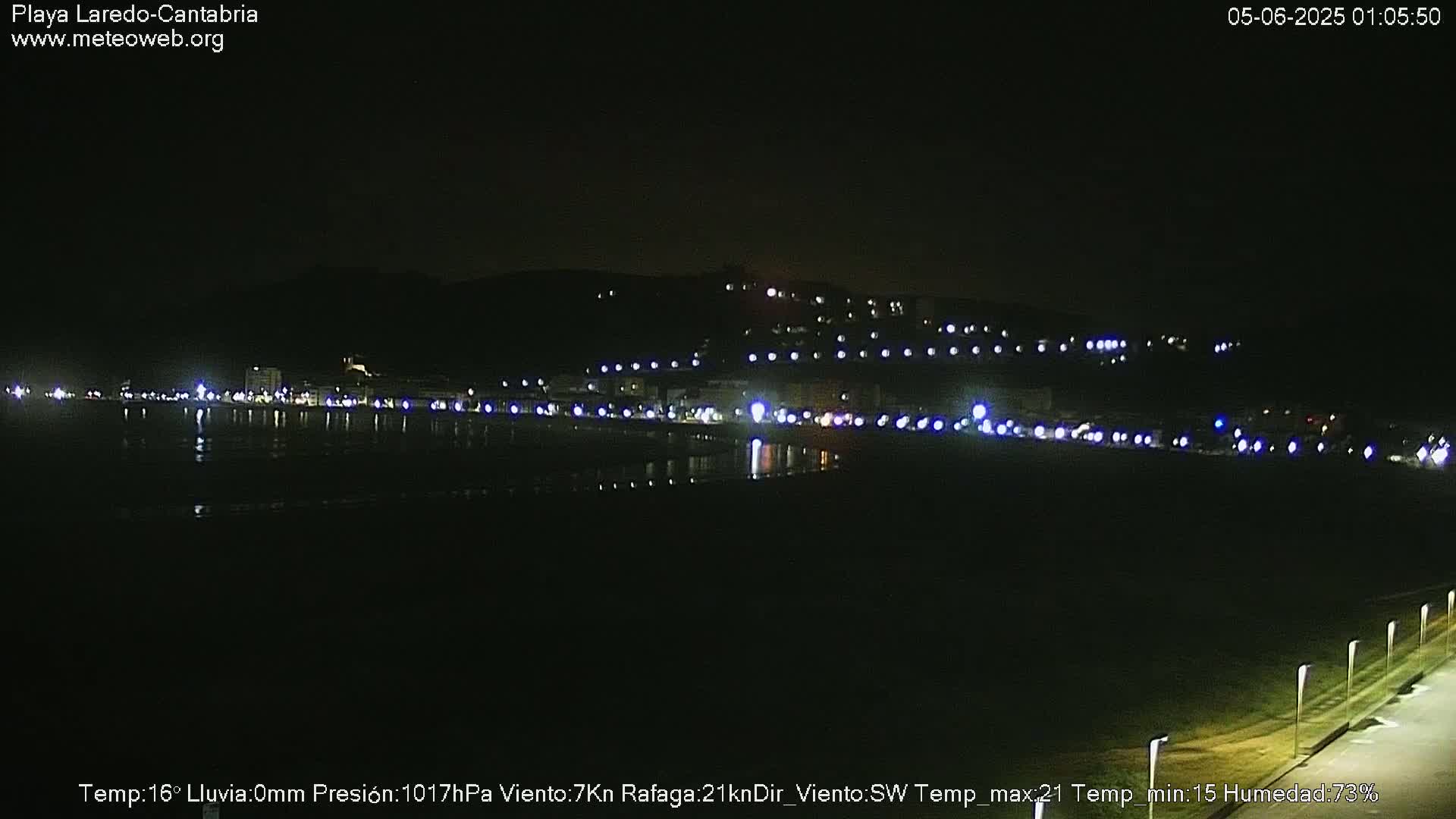 A nighttime view of a coastal town with lights lining a beach and waterfront, under a clear, dark sky.