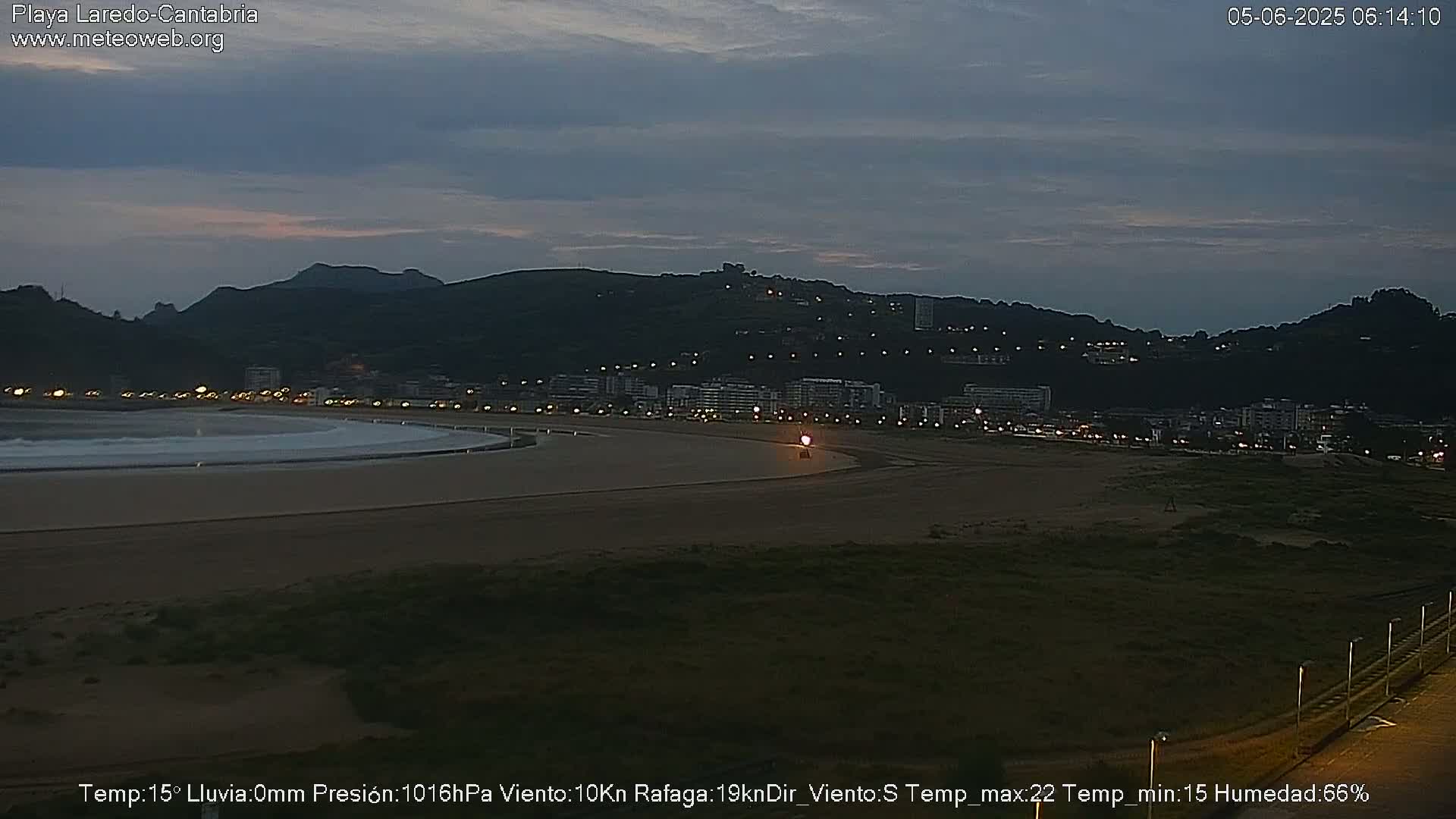 A dark, coastal town is visible at twilight under a cloudy sky, with a wide sandy beach in the foreground.
