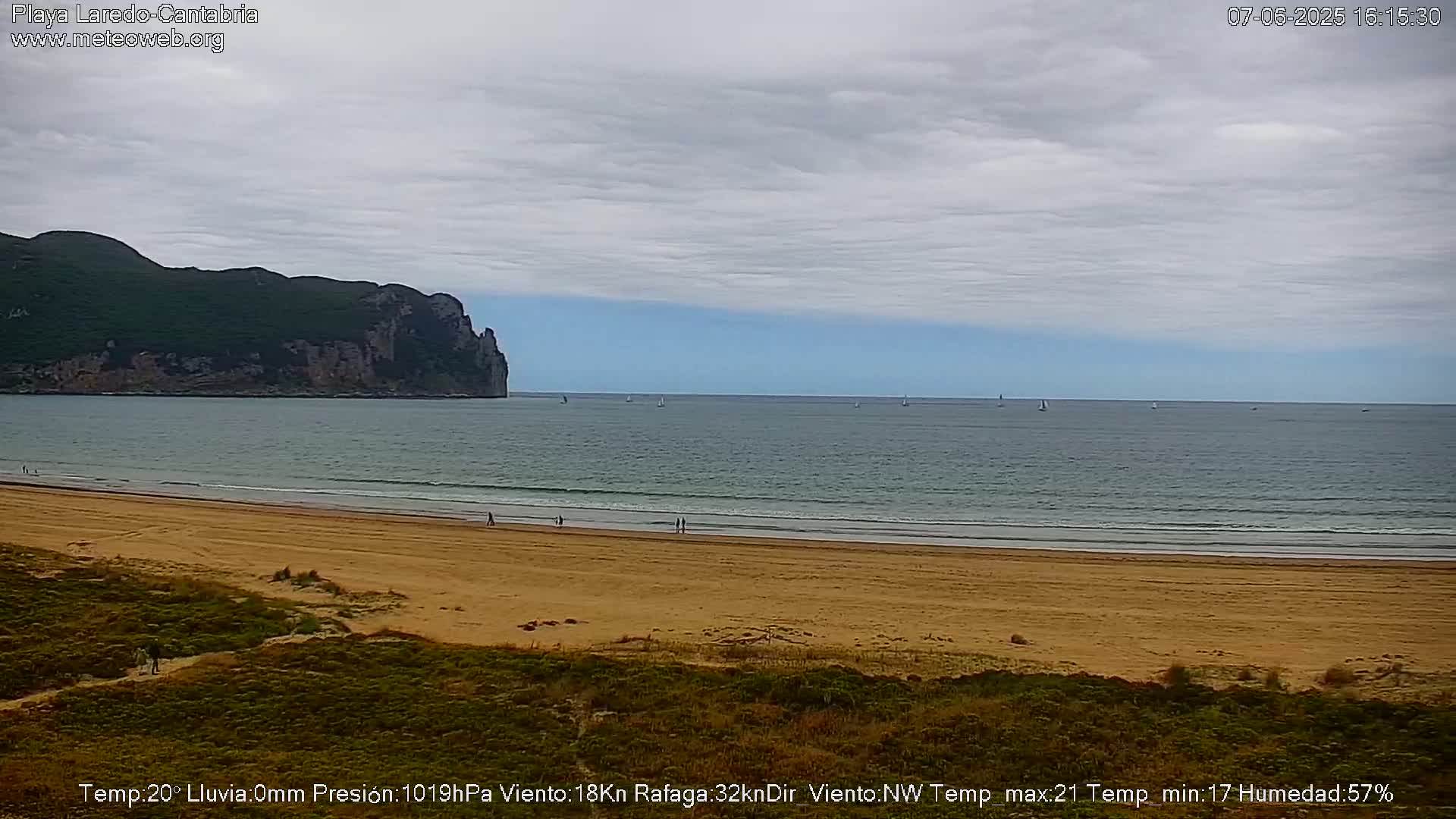 A sandy beach stretches toward a calm ocean under a mostly cloudy sky, with a dark, rocky headland visible in the distance and several sailboats on the water.