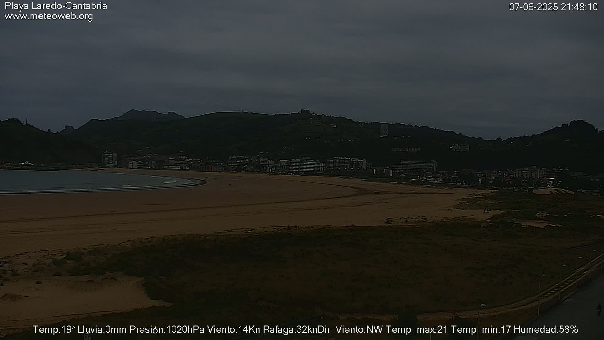 A dark, overcast beach scene shows a sandy shoreline curving around a calm bay, backed by a town nestled against dark hills.