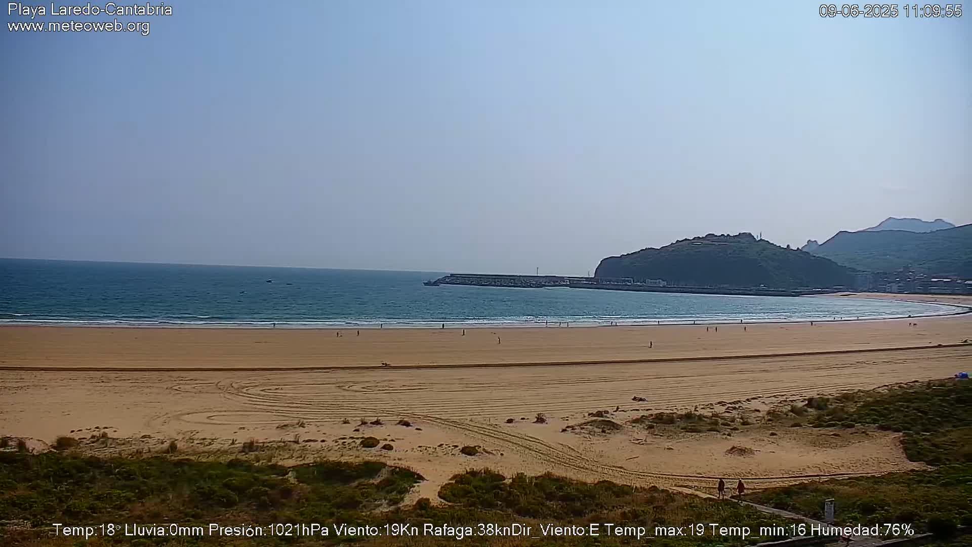 A long, sandy beach stretches out to a calm ocean under a clear, light blue sky, with a small town and hills visible in the distance.