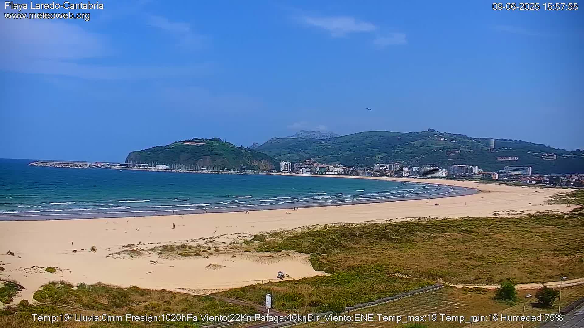 A long sandy beach stretches alongside calm ocean waves under a clear blue sky, backed by green hills and a town.