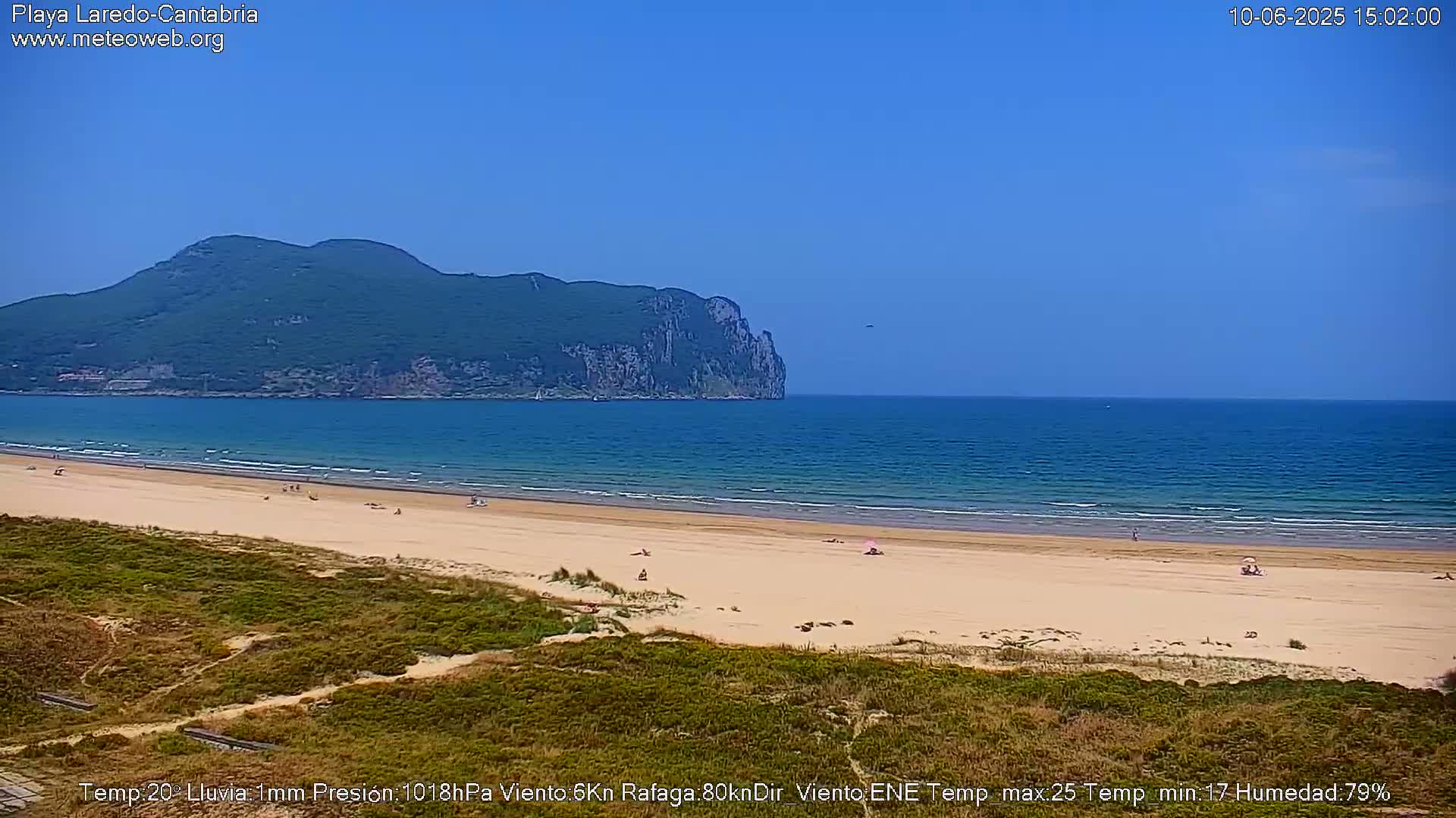 A sandy beach with a few people on it stretches along a calm ocean under a clear blue sky, with a large, verdant mountain visible in the distance.