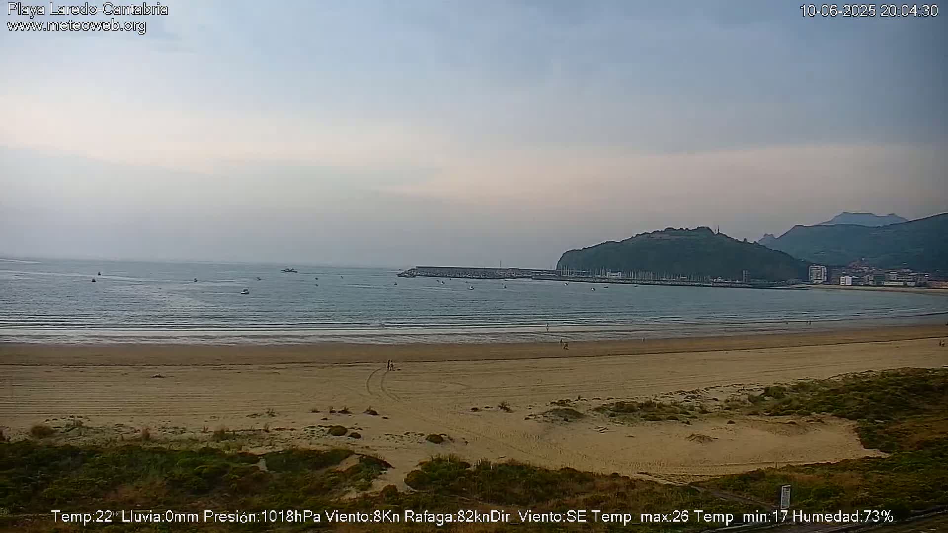 A sandy beach stretches to a calm sea under a mostly clear, light gray sky, with several small boats visible on the water and a hill in the background.