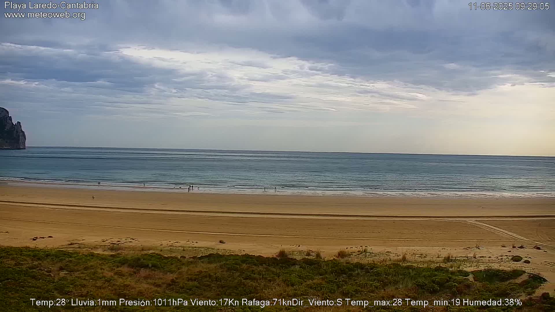 A sandy beach stretches to a calm ocean under a mostly cloudy sky.