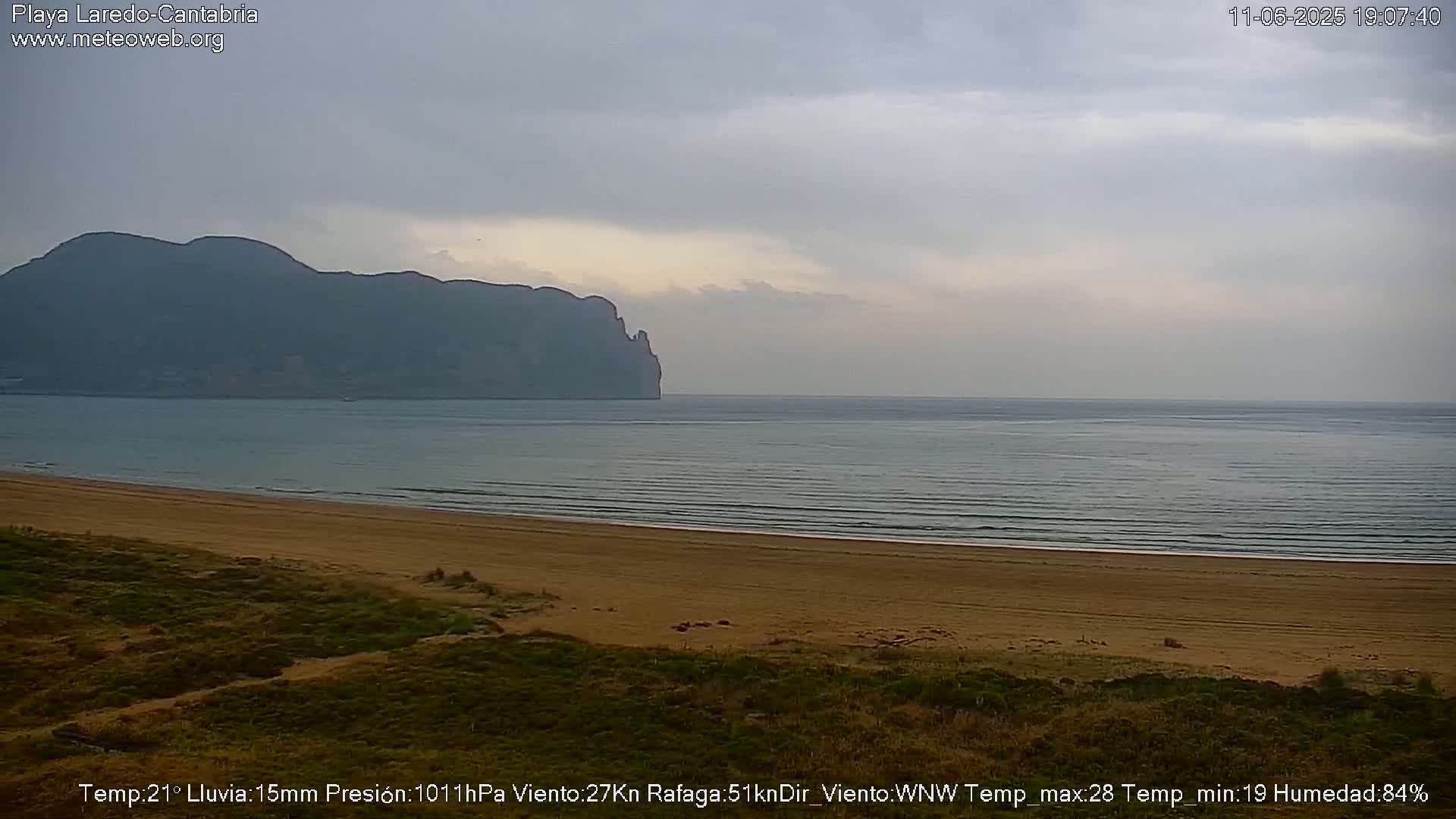 A sandy beach stretches to a calm sea under an overcast sky, with a distant mountain range visible.