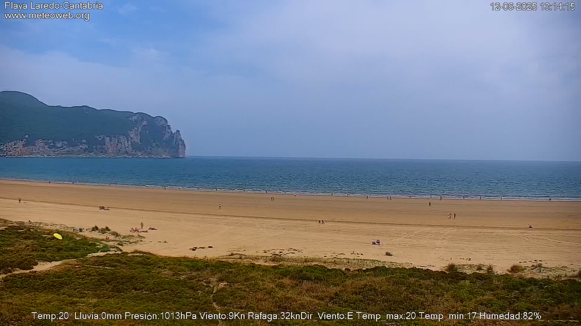 A sandy beach with scattered people stretches out to a calm ocean under a mostly clear sky, backed by a large, green-covered cliff.