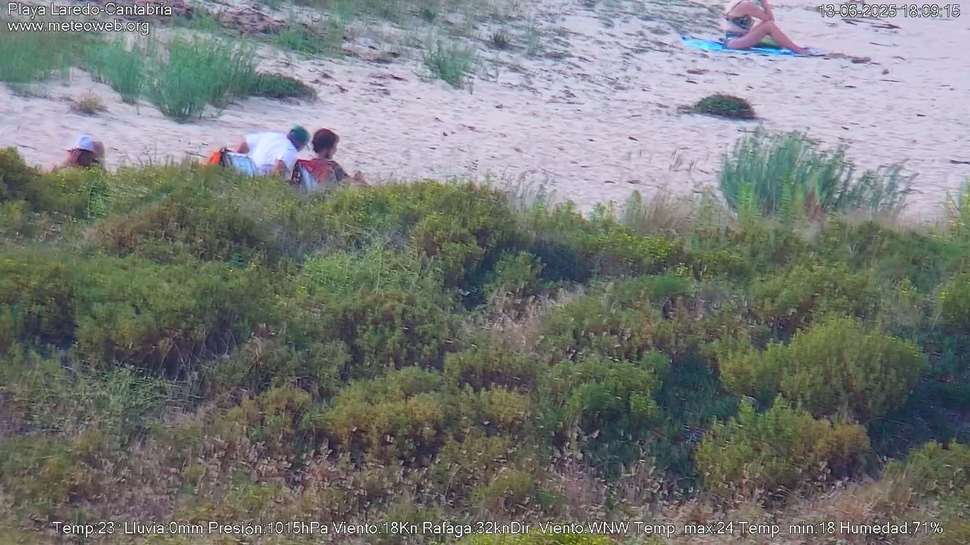 A sandy beach with several people sitting on the sand and one person lying on a towel, viewed from a slightly elevated vantage point overlooking low-lying green vegetation.