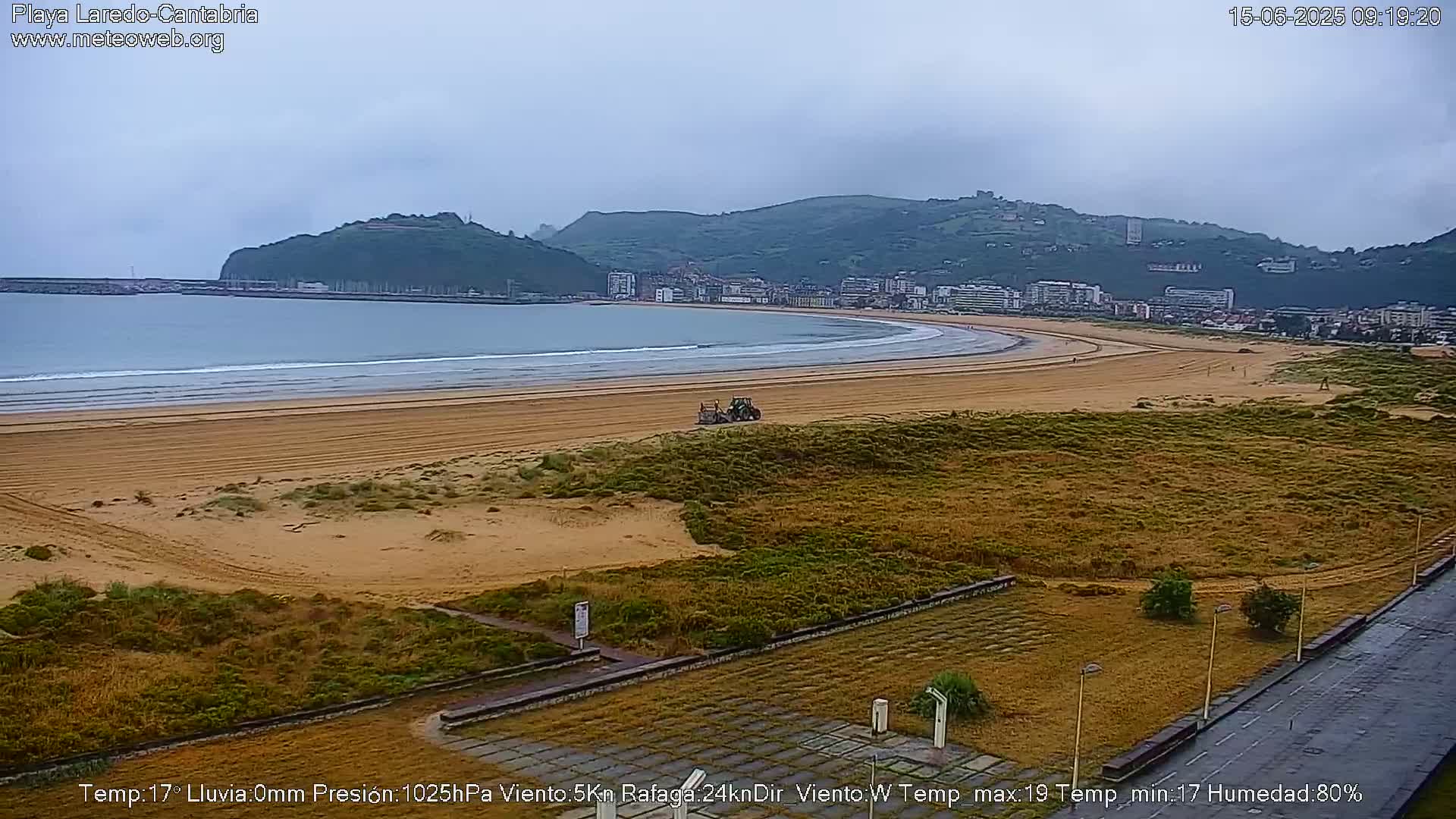 A tractor sits on a sandy beach overlooking a town nestled against a hillside under an overcast sky.