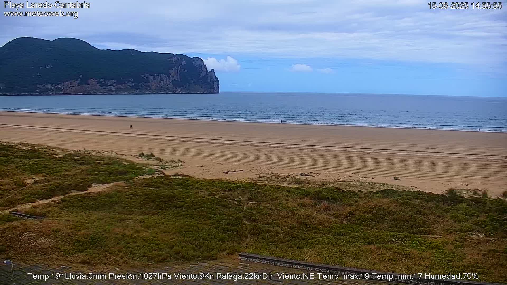 A sandy beach stretches towards a calm ocean under a partly cloudy sky, with a large, verdant mountain visible in the distance.