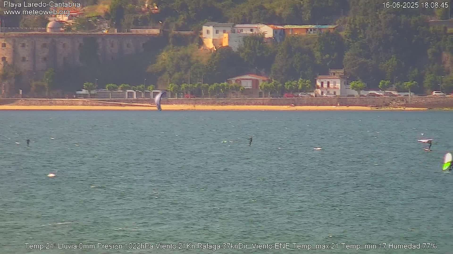 A body of water shows several people engaged in water sports, with a sandy beach and buildings in the background under sunny conditions.