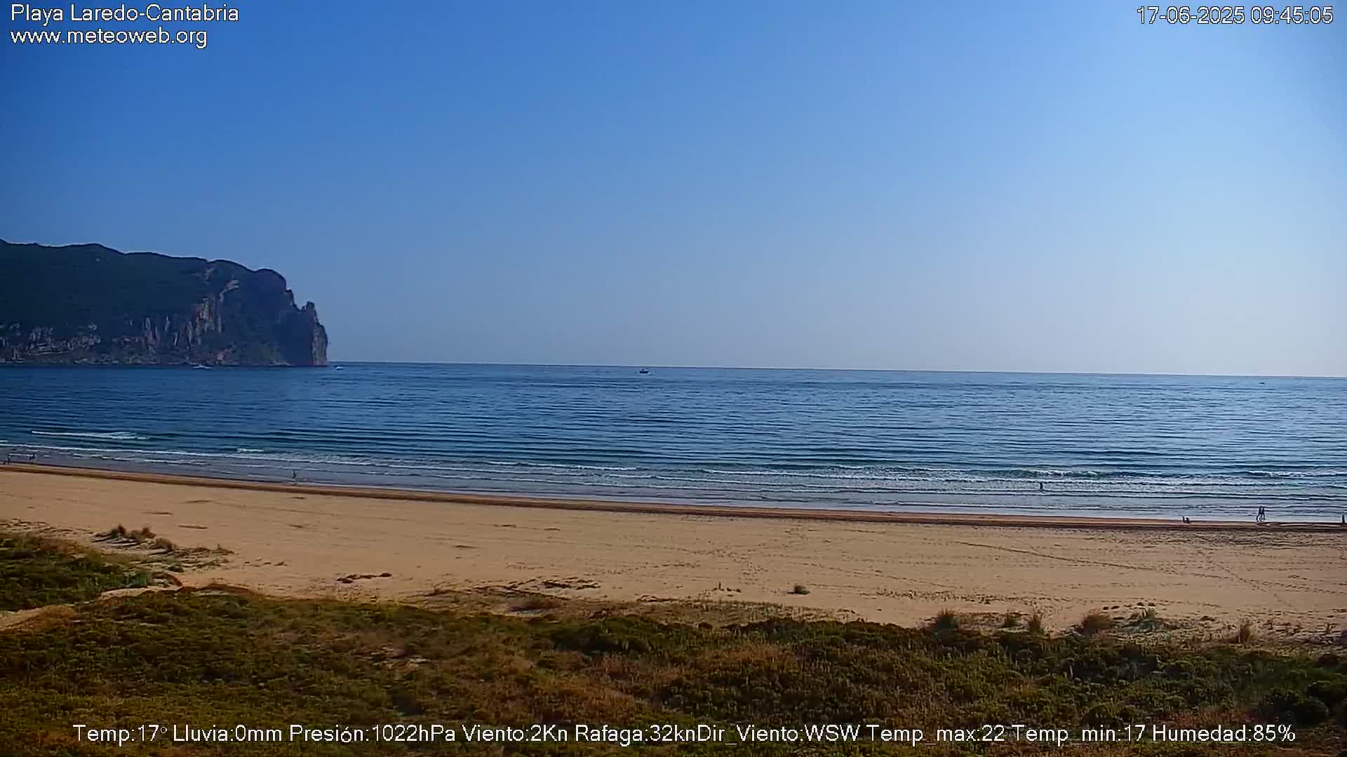 A sandy beach stretches to a calm ocean under a clear blue sky, with a large cliff visible in the distance.