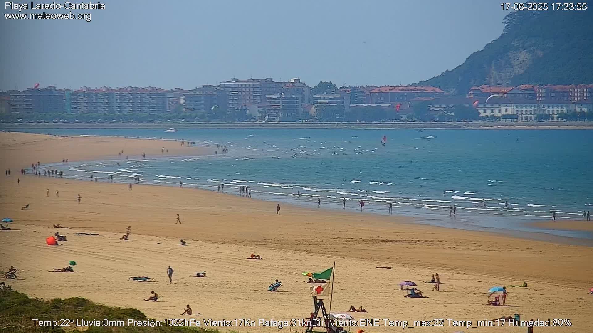A sandy beach with people sunbathing and swimming in a calm, blue ocean under a clear sky, backed by a town and a hill.