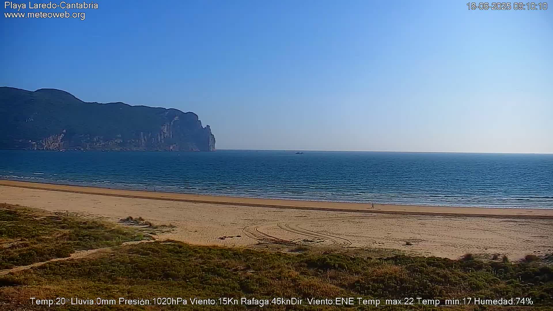 A sandy beach stretches along a calm ocean under a clear blue sky, with a large dark mountain visible in the distance.