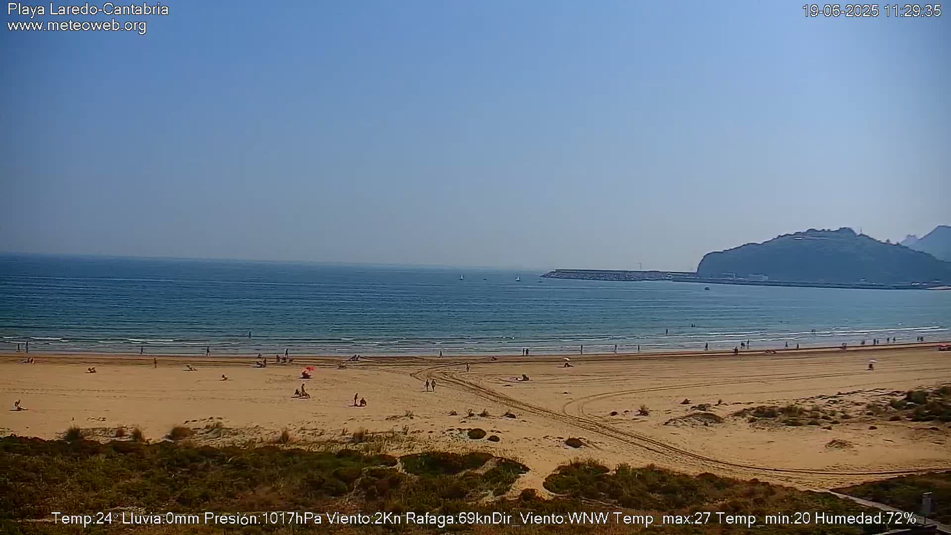A sandy beach with scattered people stretches out to a calm ocean under a clear blue sky, and a landmass is visible in the distance.