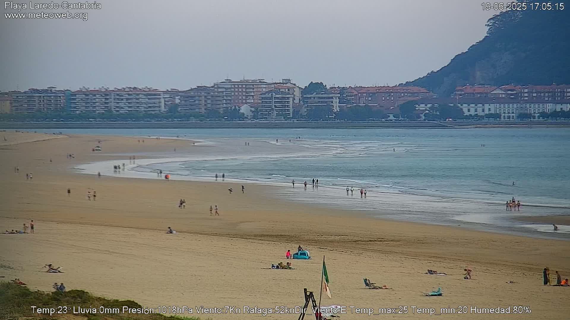 A sandy beach with scattered people on it stretches towards a calm ocean under an overcast sky, with a town and a hill visible in the background.