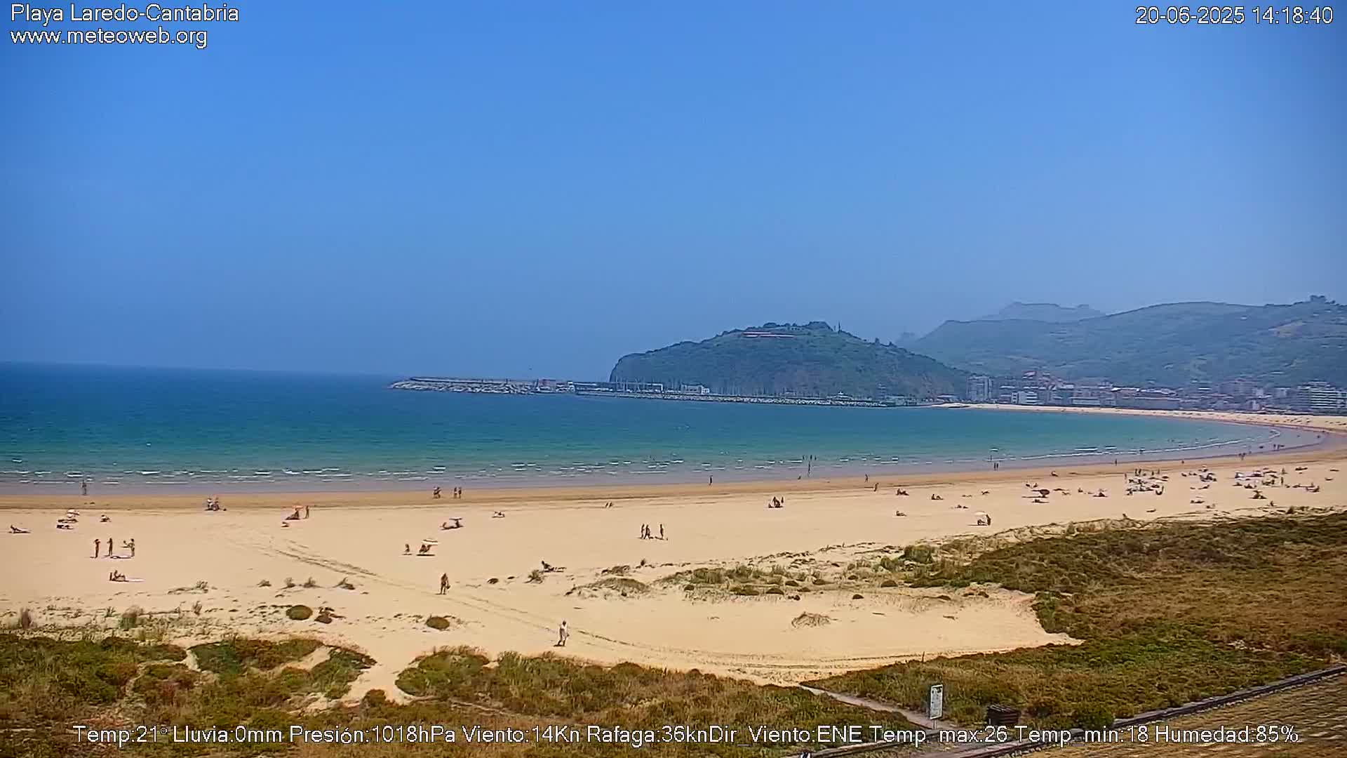 A sandy beach with scattered people stretches along a calm, teal ocean under a clear blue sky, backed by green hills and a town.