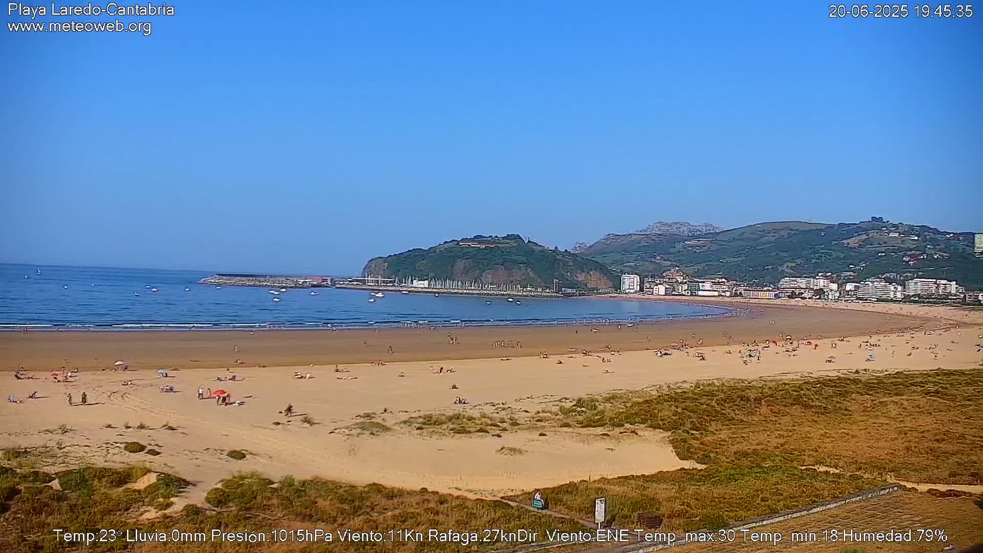 A sandy beach with scattered sunbathers stretches beneath a clear blue sky, backed by a town and a green hill.