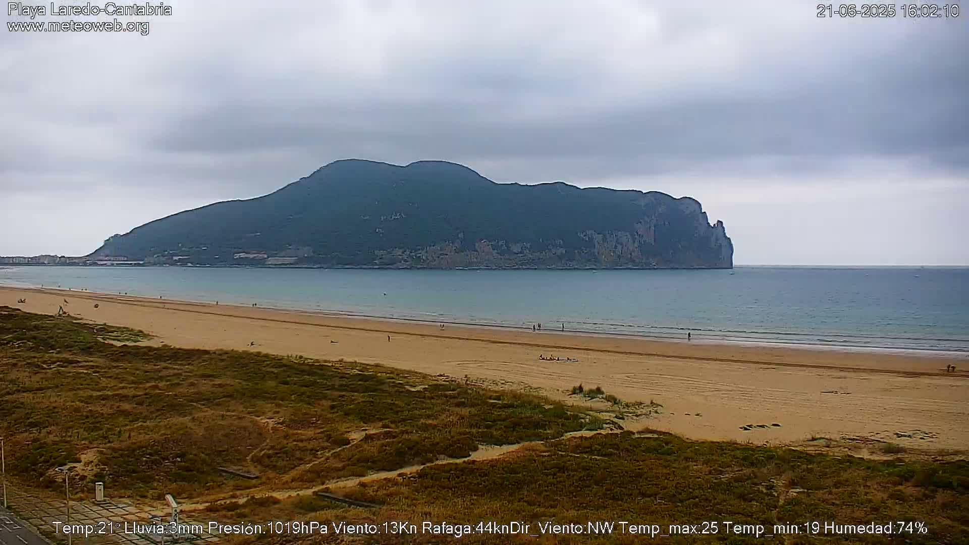 A sandy beach stretches toward a large, dark-green mountain across a calm, light-blue ocean under an overcast sky.
