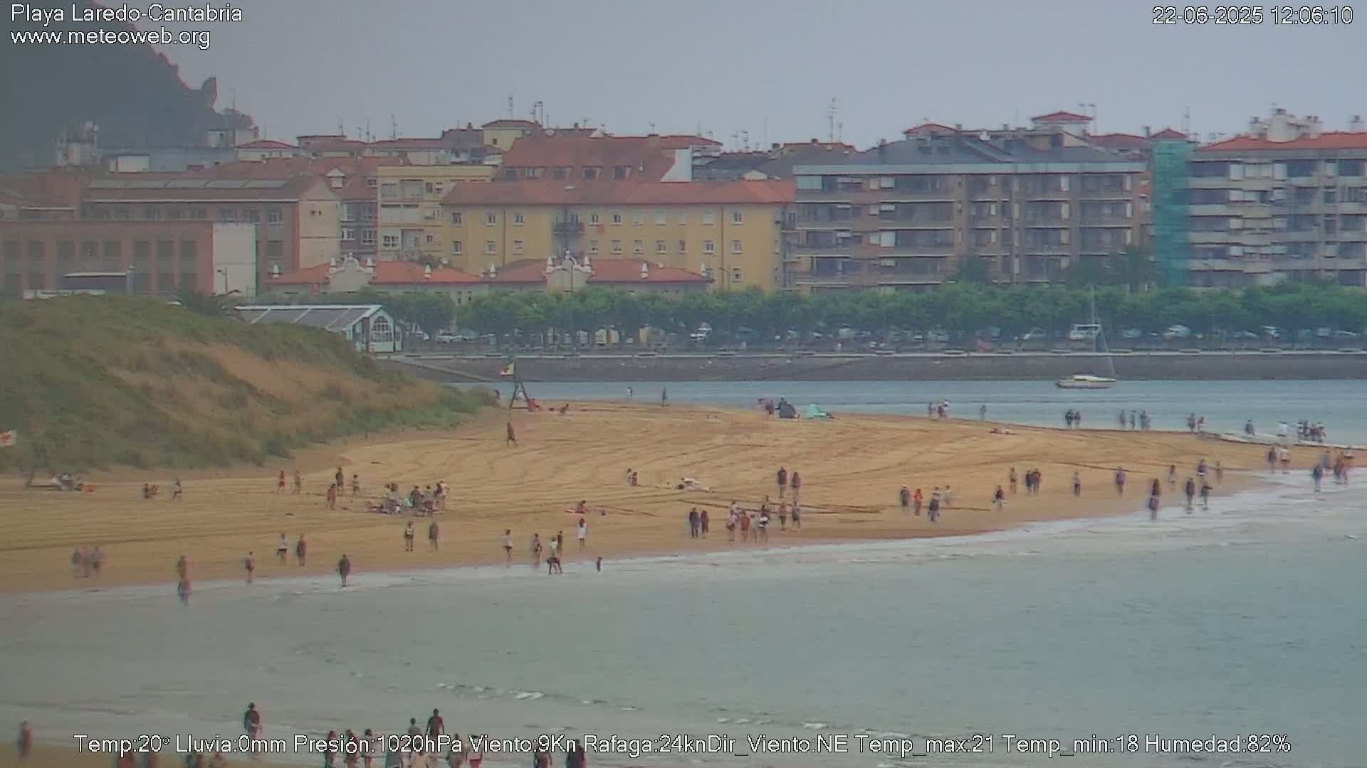 A sandy beach with many people on it is visible, extending into calm, gray water in front of a town with multistory buildings under an overcast sky.