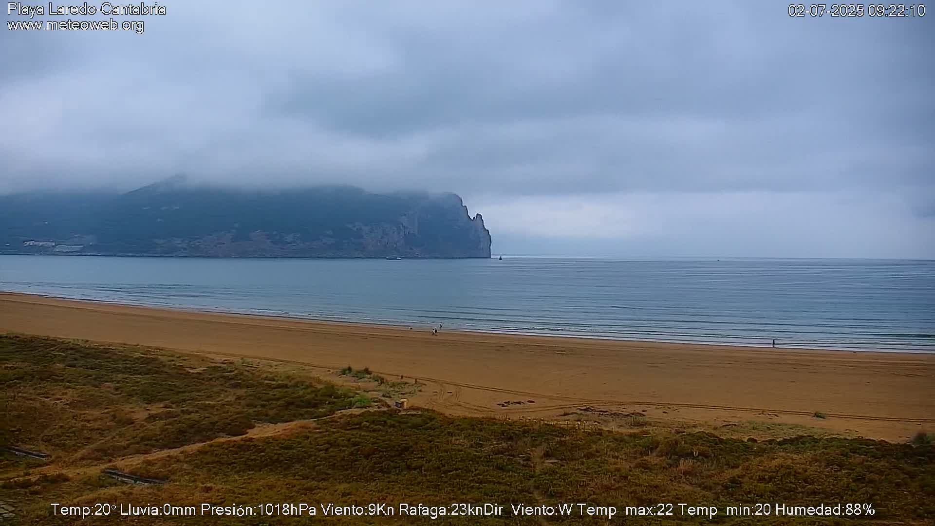 A sandy beach stretches to a calm sea under a cloudy sky, with a large, partially fog-shrouded mountain in the background.