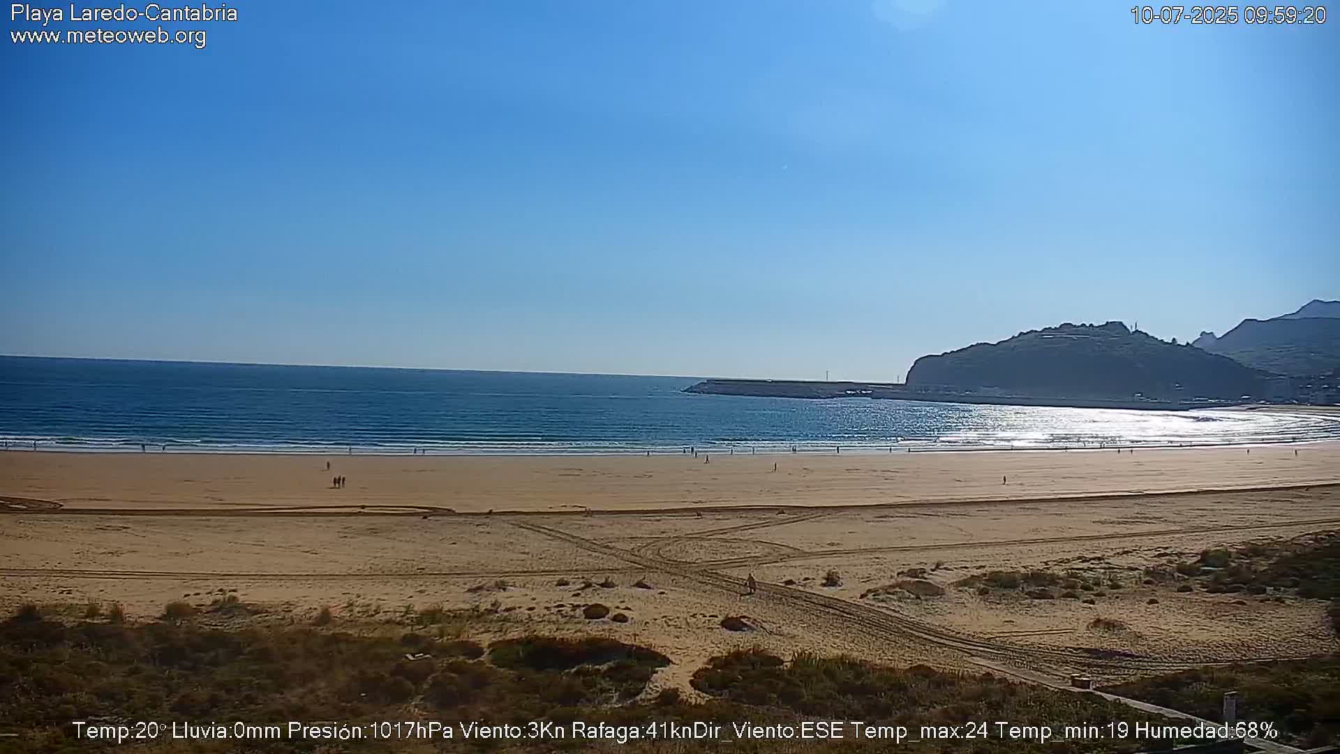 A sandy beach with a few people on it stretches out to a calm blue ocean under a clear blue sky.