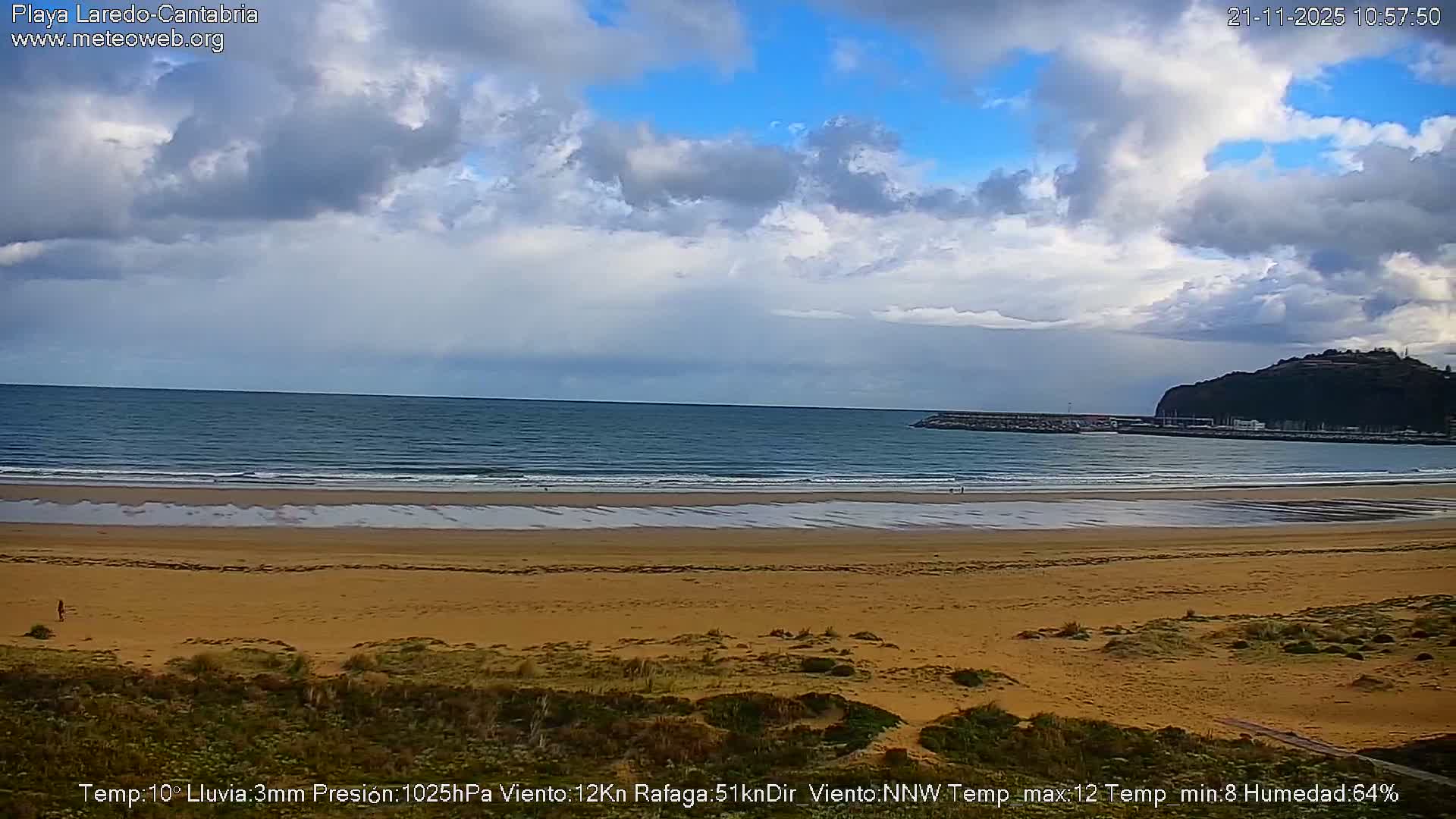 A wide sandy beach slopes towards a calm blue sea under a partly cloudy sky, with a dark, tree-covered headland and a harbor visible on the right.