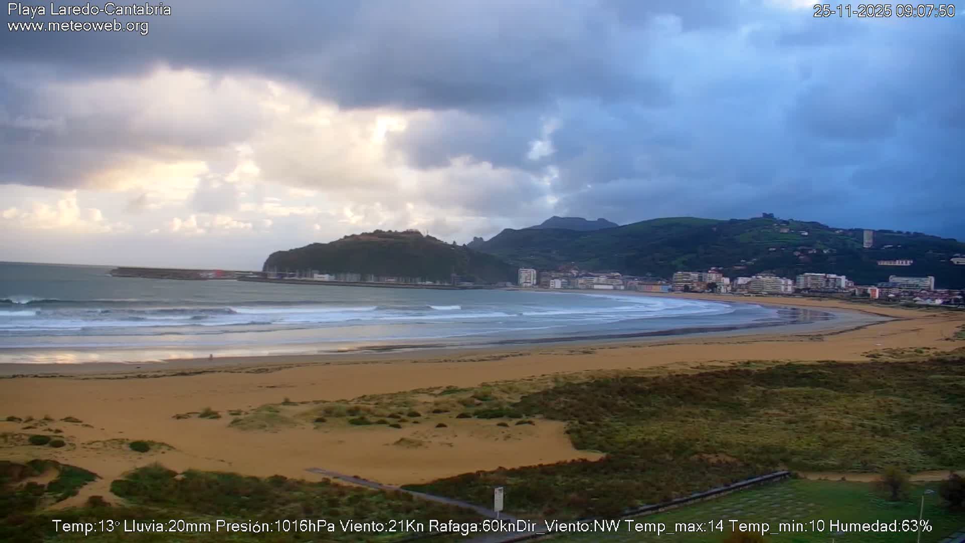 A wide sandy beach with rolling waves curves towards a coastal town nestled against verdant hills, all under a dramatic, heavily overcast sky with patches of brighter light breaking through.