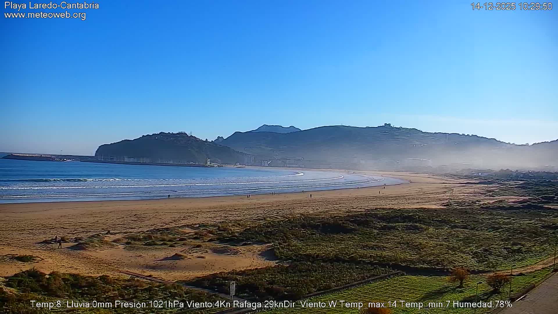 A wide shot reveals a sandy beach with gentle waves meeting a vast ocean under a partly cloudy sky with prominent grey clouds and patches of blue, stretching towards a distant headland and port on the right.