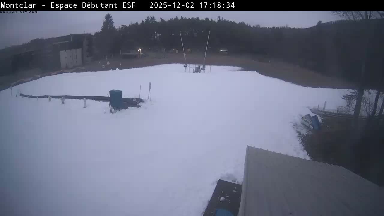 An outdoor beginner ski area is covered in snow with various equipment, distant buildings, and trees visible under a cold, overcast, and dim sky.