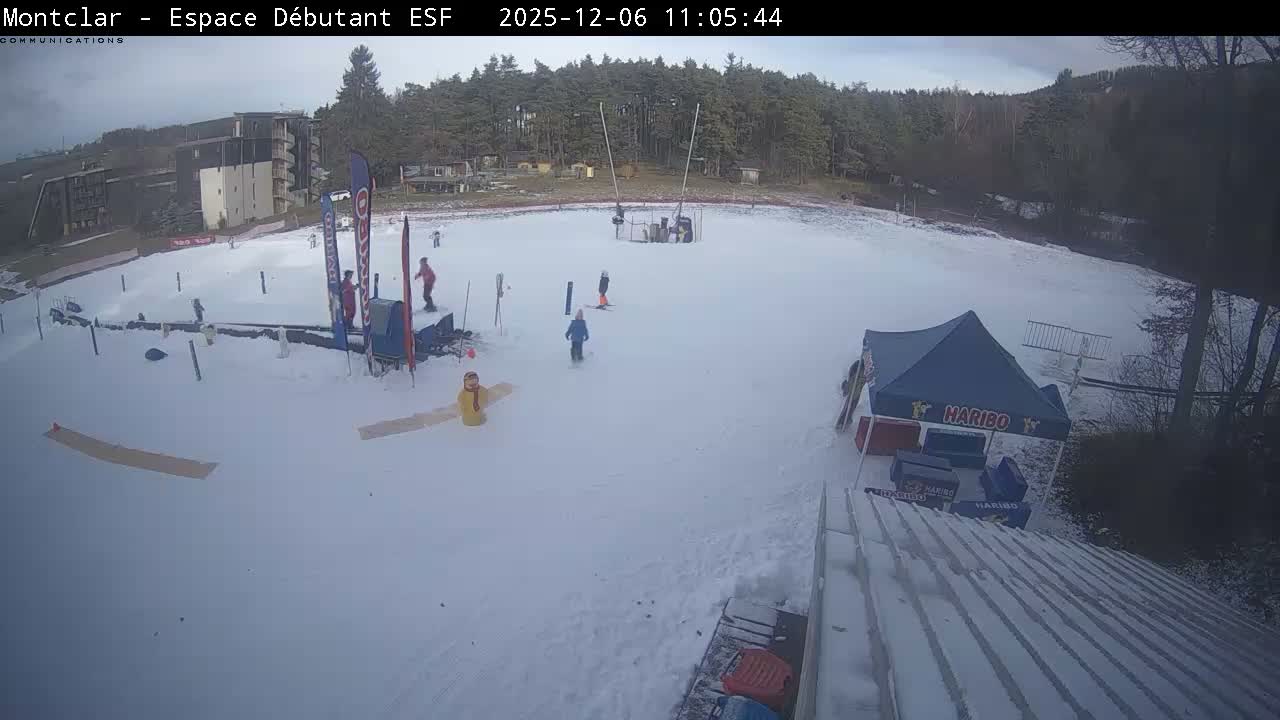 A wide shot captures a snowy ski beginner's area with several people skiing and snowboarding, bordered by buildings and a forest under an overcast sky.
