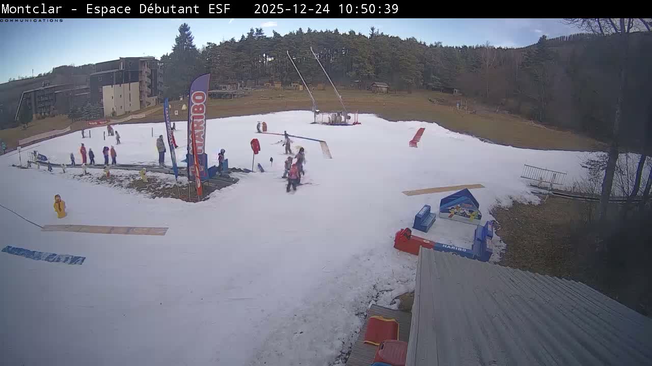 A wide shot captures a snowy ski beginner's area with several people skiing and snowboarding, bordered by buildings and a forest under an overcast sky.