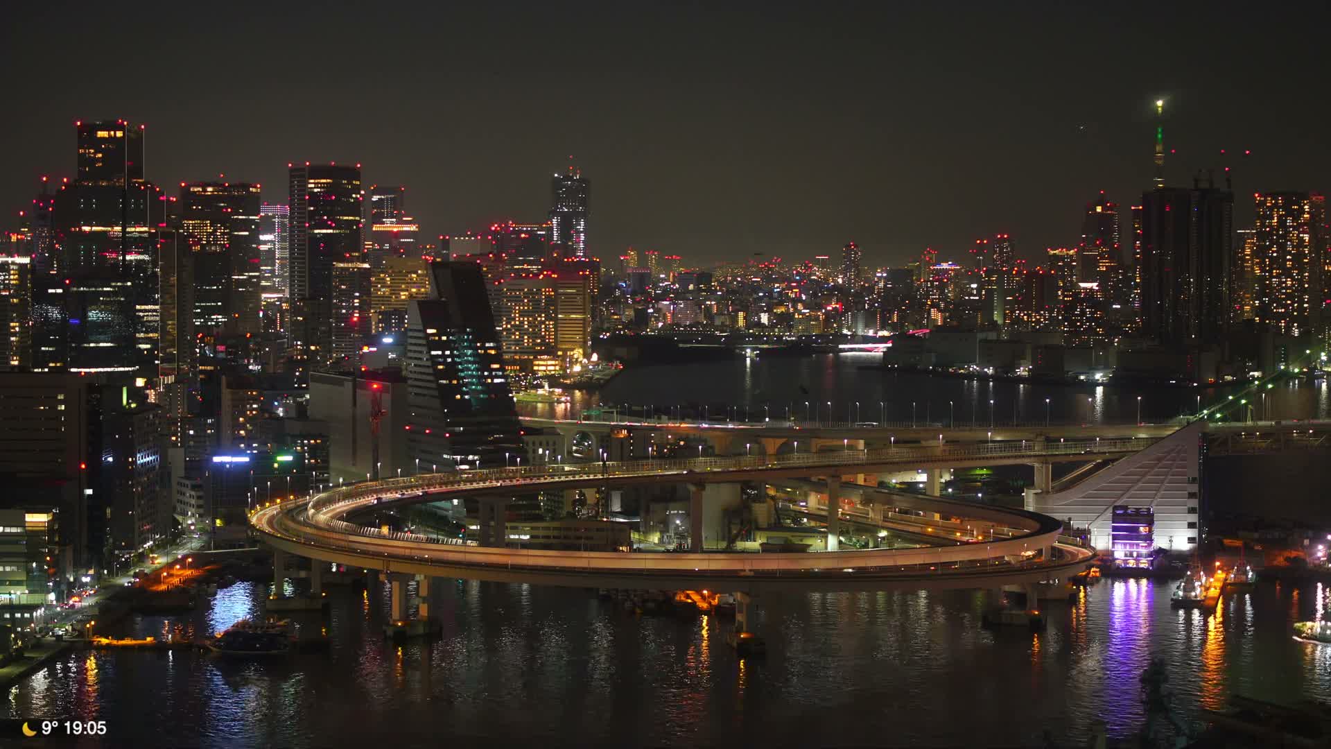 The image captures a sprawling city at night, showcasing numerous illuminated skyscrapers, complex multi-level bridges with glowing traffic winding over dark water, all beneath a clear sky.