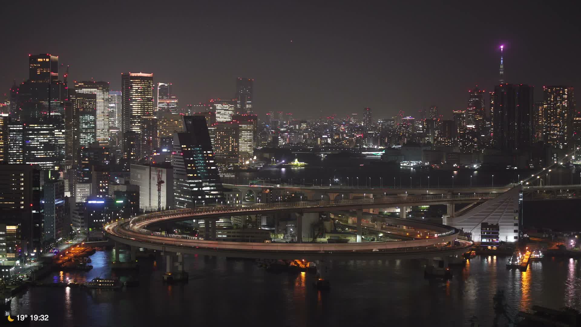 The image captures a sprawling city at night, showcasing numerous illuminated skyscrapers, complex multi-level bridges with glowing traffic winding over dark water, all beneath a clear sky.