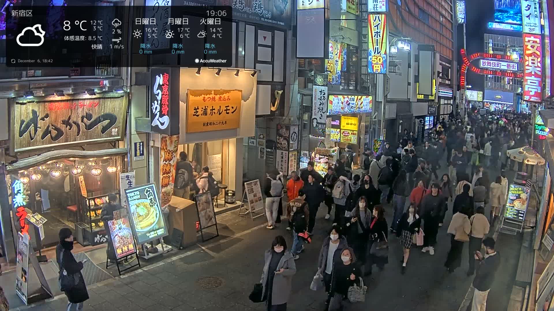 A crowded urban street at night is bustling with numerous pedestrians walking past brightly lit shops and restaurants, under cloudy skies with a temperature of 8°C.