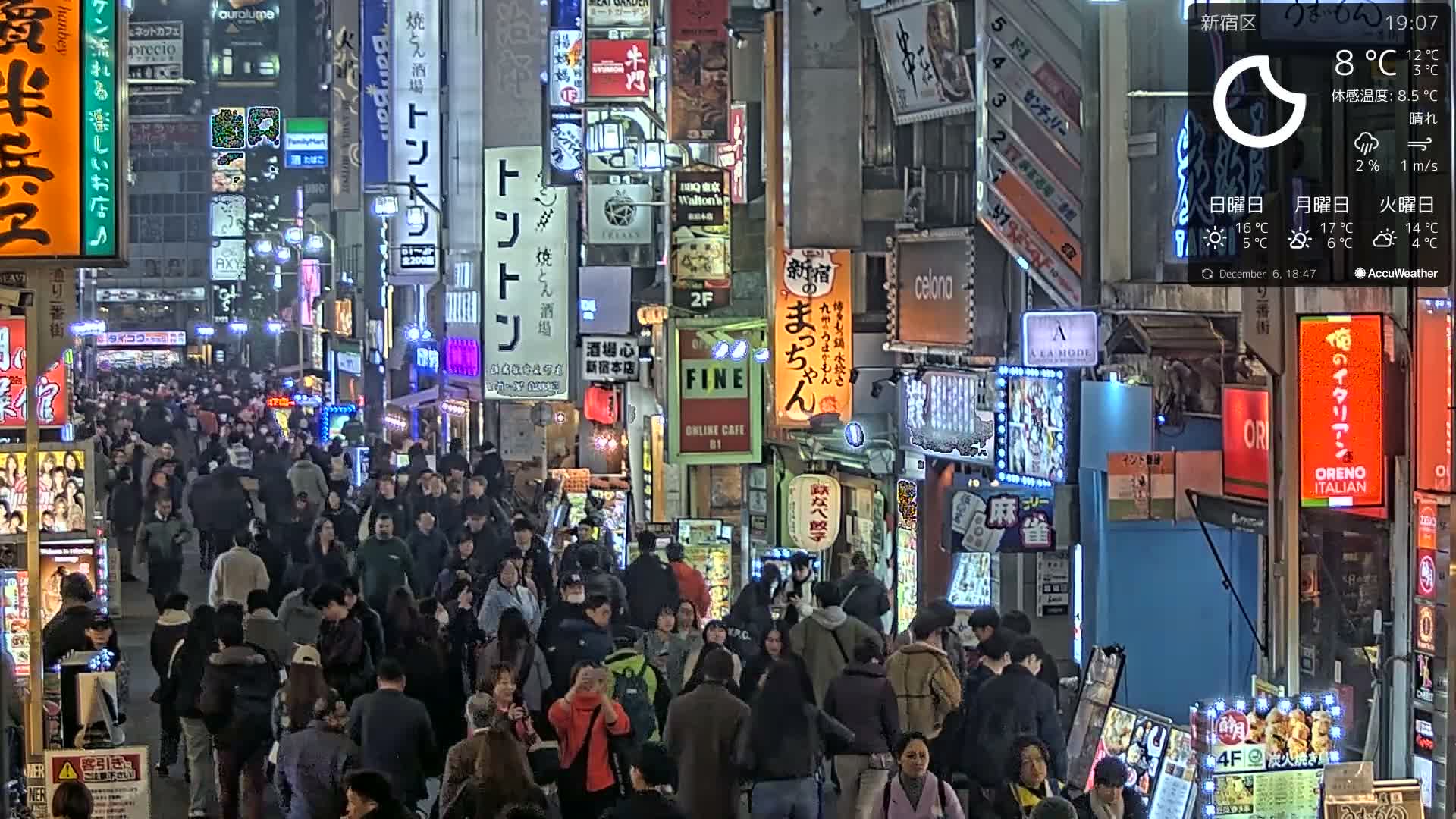 A dense crowd of people walks along a brightly lit city street at night, flanked by buildings covered in numerous colorful illuminated signs, under clear weather conditions with a temperature of 8°C.