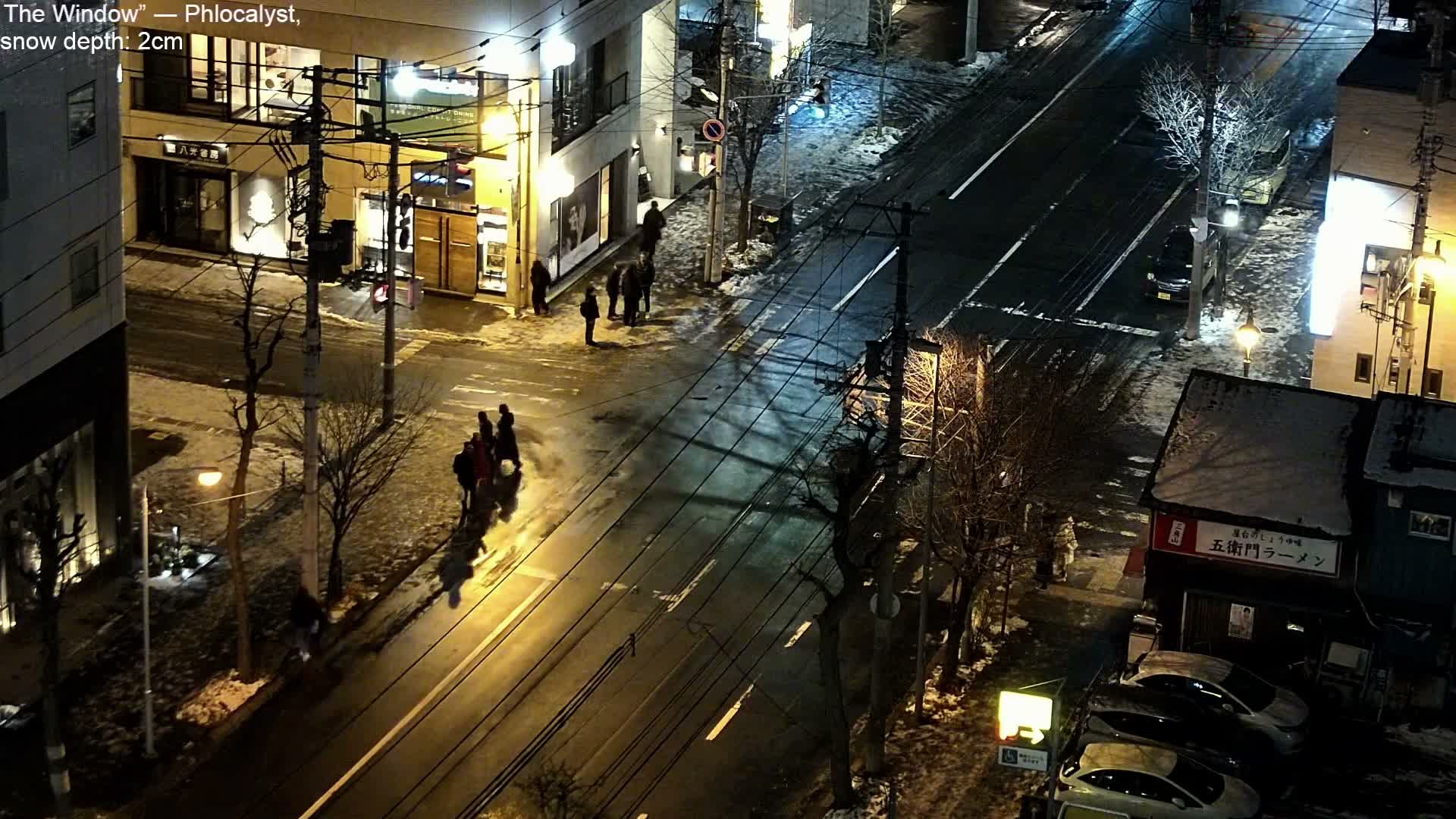 An elevated view captures a cold, snowy urban street scene at night, featuring brightly lit buildings, a wet road with a few cars, and groups of pedestrians walking along the sidewalks.