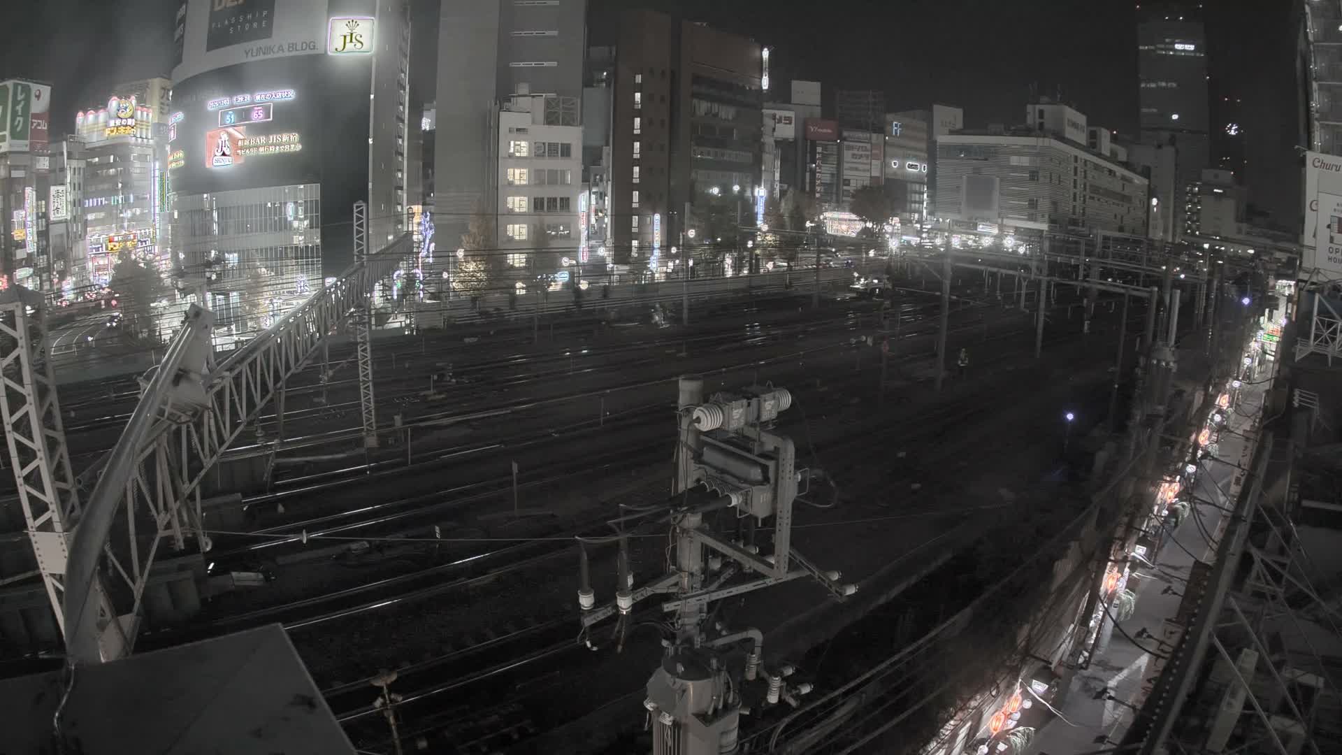 An urban landscape at night features numerous train tracks, intricate overhead power lines, and brightly lit city buildings under a clear sky.