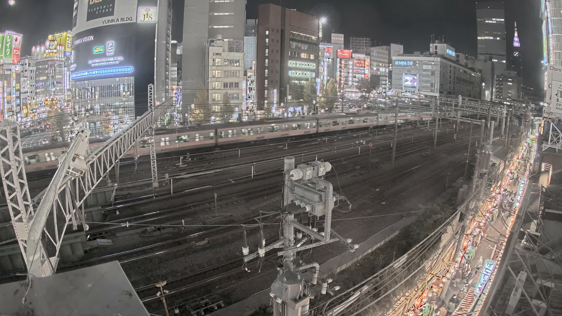 A blurred train moves along multiple tracks through a brightly illuminated urban landscape at night, featuring numerous city buildings and complex railway infrastructure under a clear sky.