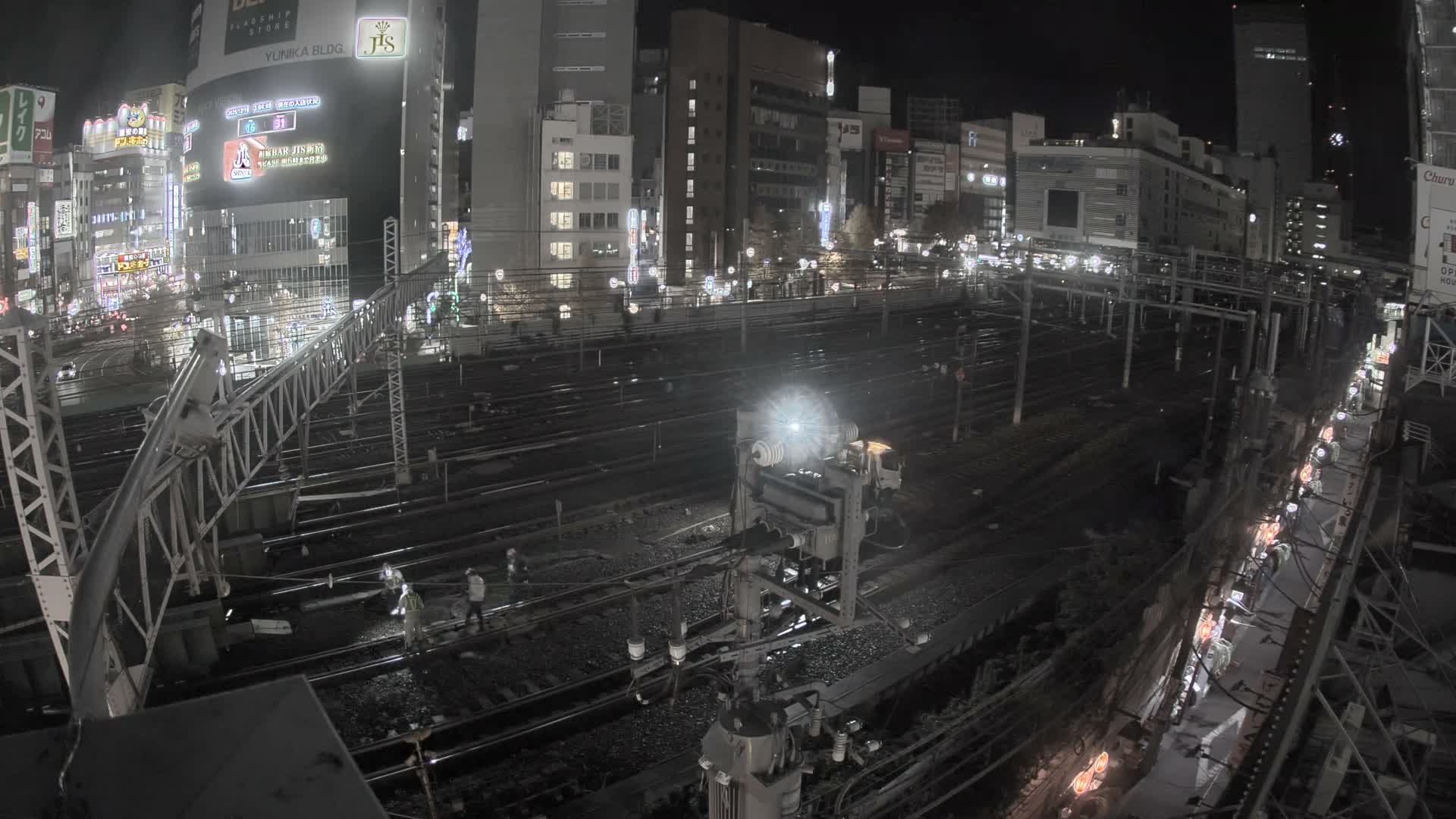 A blurred train moves along multiple tracks through a brightly illuminated urban landscape at night, featuring numerous city buildings and complex railway infrastructure under a clear sky.