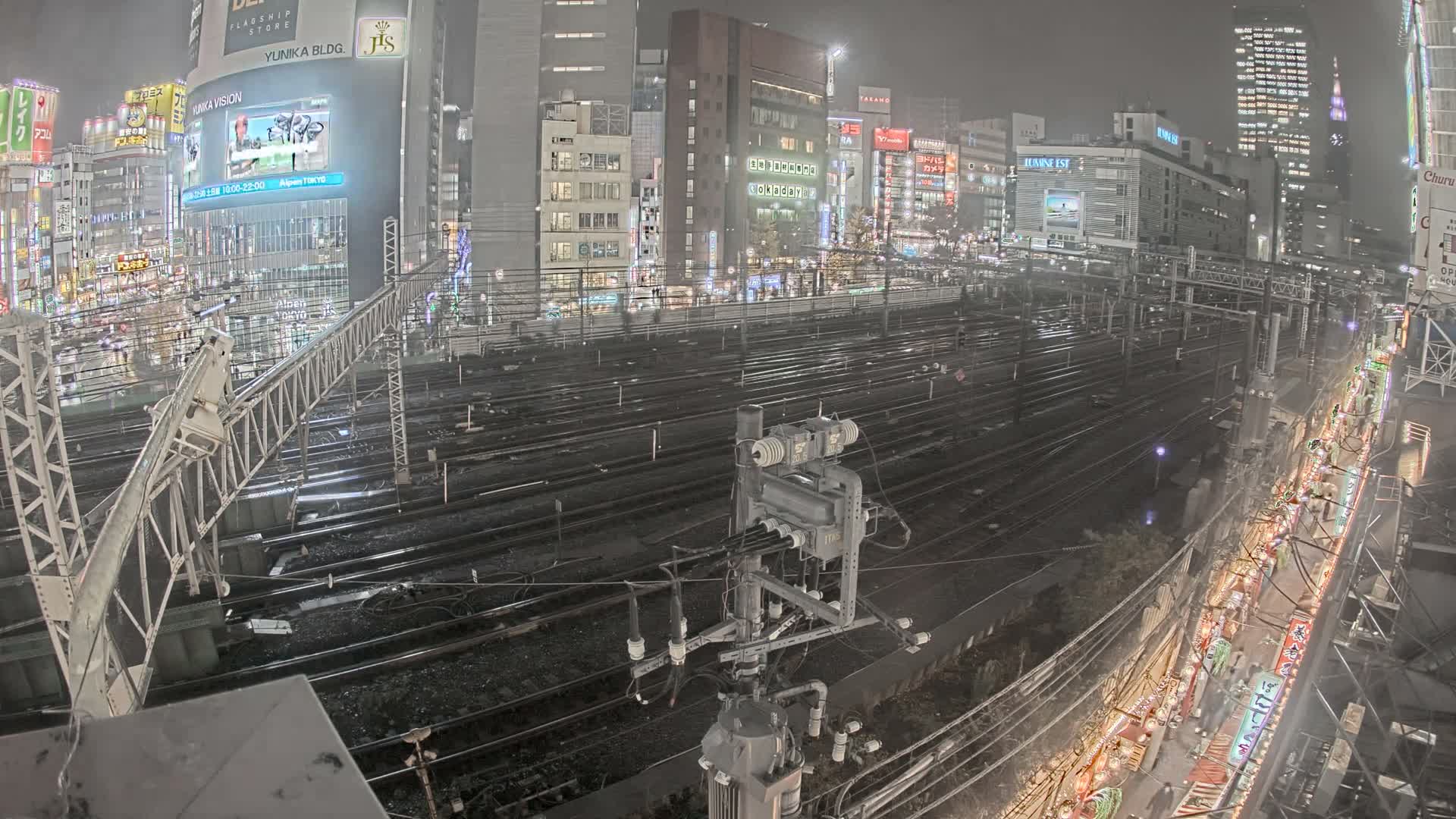 A blurred train moves along multiple tracks through a brightly illuminated urban landscape at night, featuring numerous city buildings and complex railway infrastructure under a clear sky.
