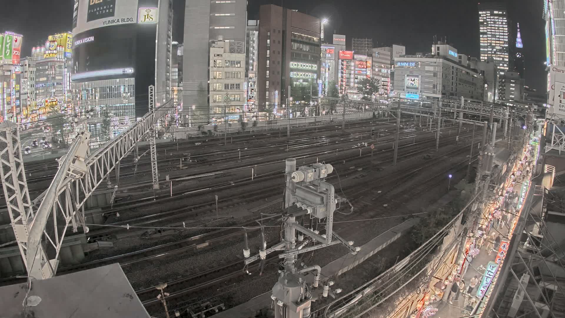 A blurred train moves along multiple tracks through a brightly illuminated urban landscape at night, featuring numerous city buildings and complex railway infrastructure under a clear sky.