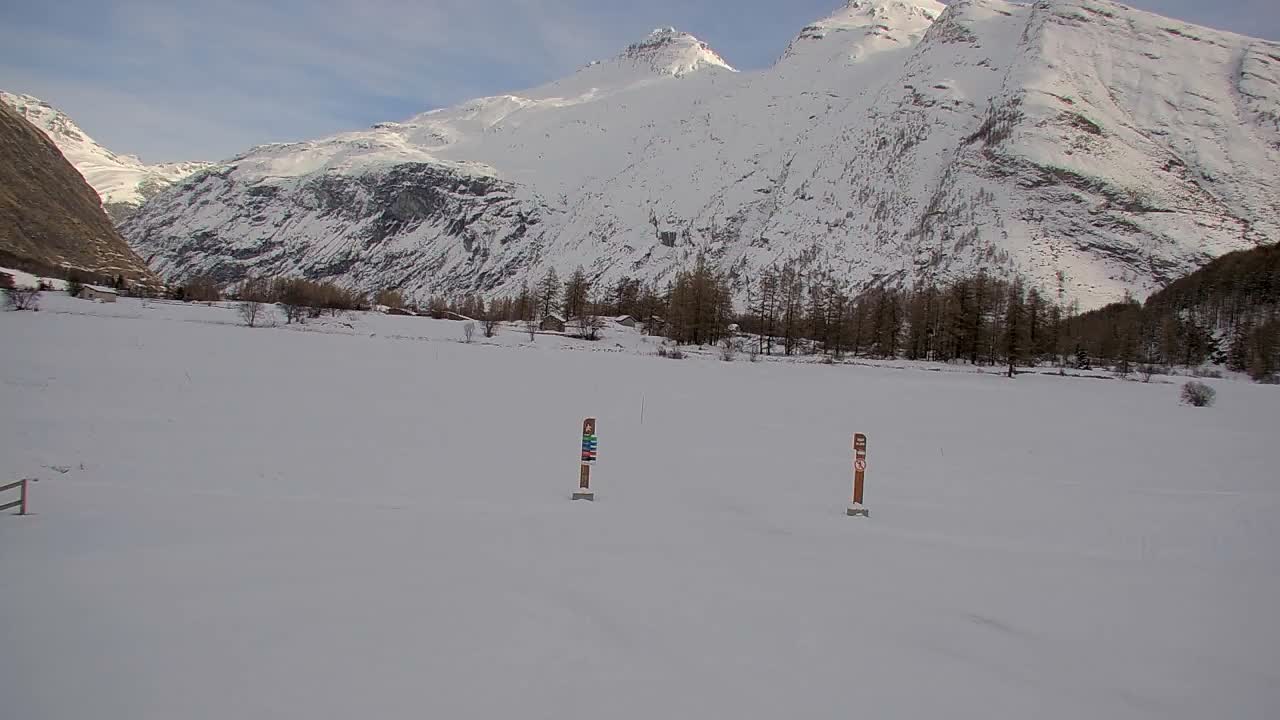 A vast snow-covered valley floor with two markers in a wide field leads to a line of evergreen trees and scattered buildings at the base of towering, snow-capped mountains under a clear to partly cloudy sky.