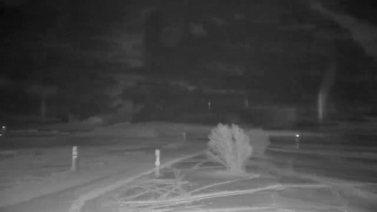 A vast snow-covered valley floor with two markers in a wide field leads to a line of evergreen trees and scattered buildings at the base of towering, snow-capped mountains under a clear to partly cloudy sky.