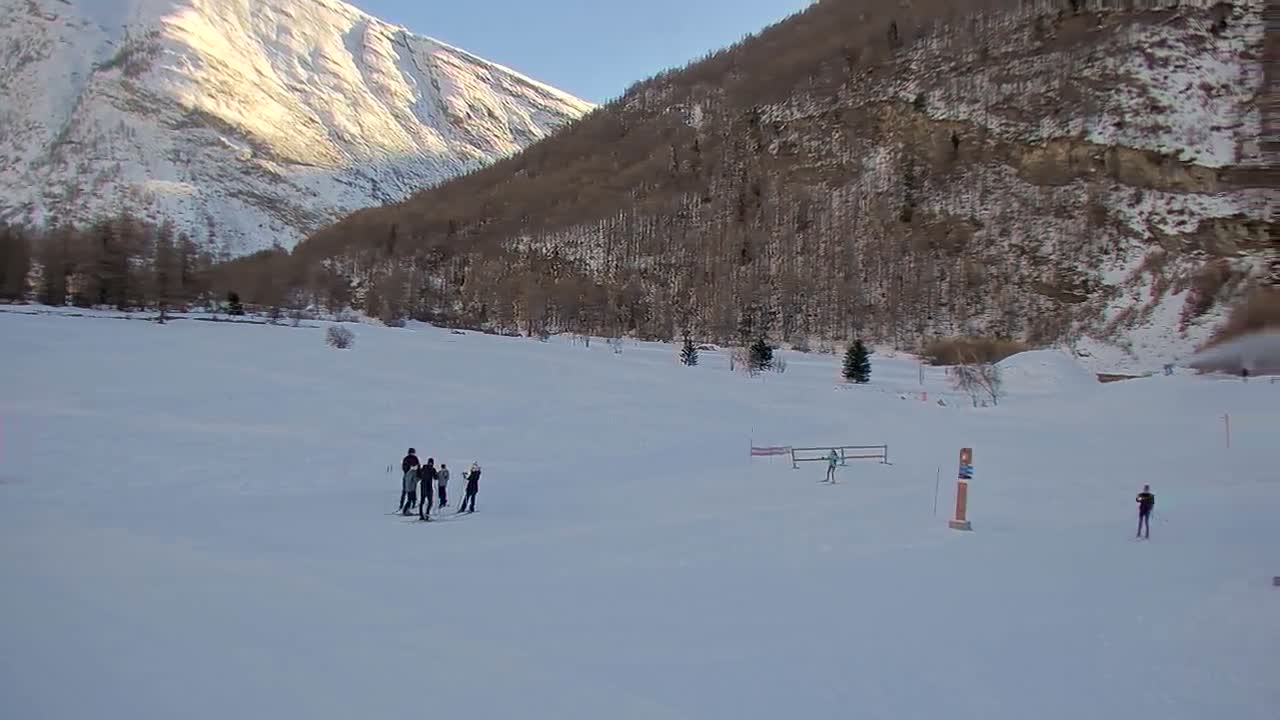 A vast snow-covered valley floor with two markers in a wide field leads to a line of evergreen trees and scattered buildings at the base of towering, snow-capped mountains under a clear to partly cloudy sky.