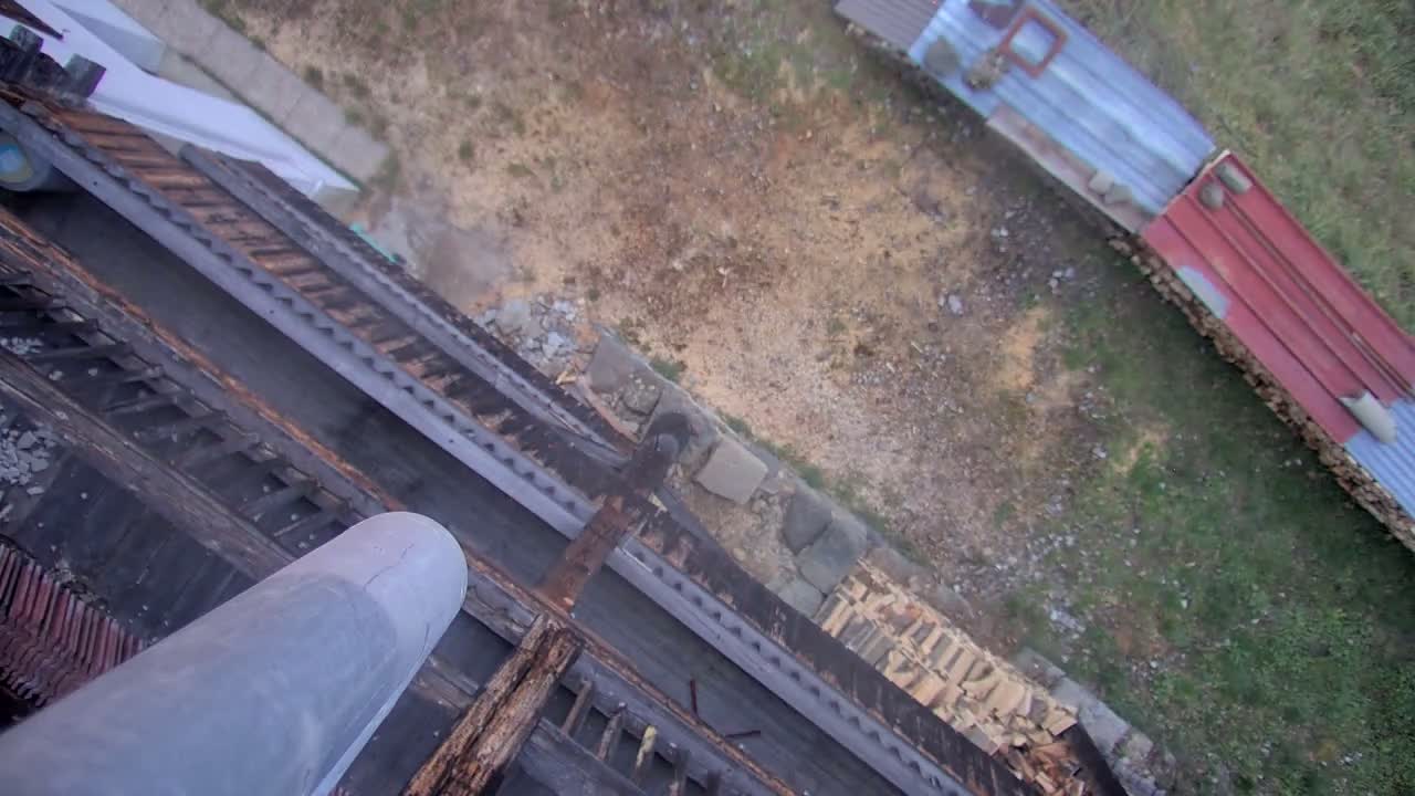 The image displays an overhead view looking down a large grey pipe onto an old wooden roof structure, beyond which lies a patchy dirt and grass yard with a long corrugated metal shed roof in the distance, under a clear or overcast daytime sky.